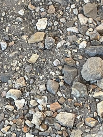 The image shows a close-up, overhead view of a gravelly surface, likely a path or ground covering in Torquay, United Kingdom. The surface is composed of numerous small to medium-sized stones, varying in color from light grey and white to darker grey and brown. Some larger, rougher stones are also present, with irregular shapes and textures. The gravel appears to be dry and loose, with some finer dirt or sand interspersed among the stones. There are no discernible people, animals, or man-made objects visible in the frame. The environment suggests an outdoor setting, possibly a garden, park, or unpaved area within the city.