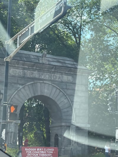 The image shows a view of the Camp Randall Memorial Arch in Madison, Wisconsin, United States. The arch is made of gray stone and has the words "CAMP RANDALL" inscribed on it. A gargoyle-like sculpture is visible on the left side of the arch. A traffic signal with a lighted pedestrian crossing symbol is situated near the arch. There is a sign that reads "BADGER WAY CLOSED FOR CONSTRUCTION DETOUR THROUGH ENGINEERING DRIVE." The surrounding environment includes trees and greenery. The image seems to be taken from inside a vehicle, as reflections and parts of the car structure are visible.