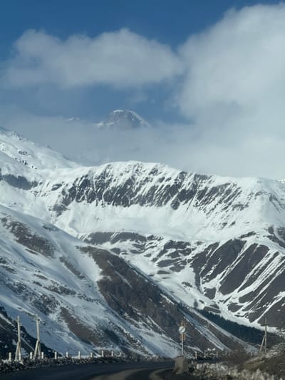 This image depicts a snow-covered mountainous landscape in the city of Kanobi, Georgia, under a partly cloudy blue sky. A paved, dark road curves through the lower foreground, extending from the lower-left towards the center-right. No individuals or vehicles are present.
The sky in the upper portion is clear blue, transitioning to white, billowy clouds that partially obscure the upper reaches of the mountains. A prominent, snow-capped peak is visible in the distant upper center, partially shrouded by these clouds.
Mid-ground and foreground mountains are heavily blanketed in white snow, revealing patches of dark, exposed rock and earth, especially on steeper slopes where snow cover is intermittent or sparse. Darker, possibly coniferous vegetation is visible in concentrated areas on the lower slopes in the distant right.
Along the left edge of the road, a series of light-colored, vertical posts, potentially guardrail supports or snow poles, are visible, accompanied by a low barrier of stone or concrete. On the right side of the road, a round traffic sign with a red border is mounted on a pole anchored in a concrete or rock base. Further right, a partial fence structure is discernible. The immediate edges of the road are lined with dark, rocky material and sparse dry vegetation.