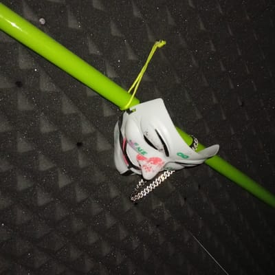The image shows a white plastic Guy Fawkes mask hanging from a green pole, which is suspended from a black ceiling covered with white triangular patterns. The mask is partially obscured by the pole, and a silver chain hangs from its chin. The mask features black eyes, a black mouth, and a red and white nose and cheek area. The mask is located in an unknown location in Biedenkopf, Germany (latitude: 50.914, longitude: 8.466). The image does not depict any people or actions.