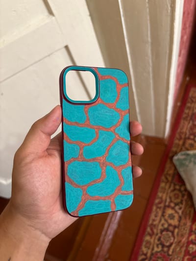 A hand holds a phone case with a teal and reddish-orange abstract pattern, resembling a giraffe or leopard print, against a backdrop of a light-colored wooden door and a red patterned rug. The phone case has a maroon border. Small smudges of blue and red paint are visible on the thumb. The scene appears to be indoors, possibly in a residential setting. The location is identified as Nukus, Uzbekistan.