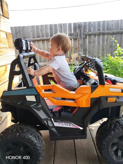 In the city of Erkhirik, Russia, a young child, likely a toddler, is seated in an orange and black toy ride-on vehicle that resembles an ATV or UTV. The child is wearing a white t-shirt and light-colored shorts. They are reaching forward with their right hand towards a set of four headlights mounted on a black roll bar above the vehicle's dashboard. The toy vehicle is positioned outdoors on a wooden deck or porch, with a weathered wooden fence in the background. Green foliage and plants are visible behind the fence, suggesting a residential or natural outdoor setting. The child's left hand is also on the roll bar. The overall scene captures a moment of a child interacting with a toy vehicle in a backyard environment.