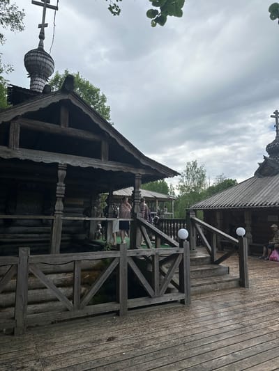 The image depicts an elevated, wooden structure in Vzglyadnevo, Russia. The structure has multiple levels connected by wooden platforms and railings. The architecture appears to be rustic, possibly of a traditional design. People are visible on the upper levels. The sky is overcast. In the lower left corner of the image is a portion of a structure topped by a cupola and a cross.