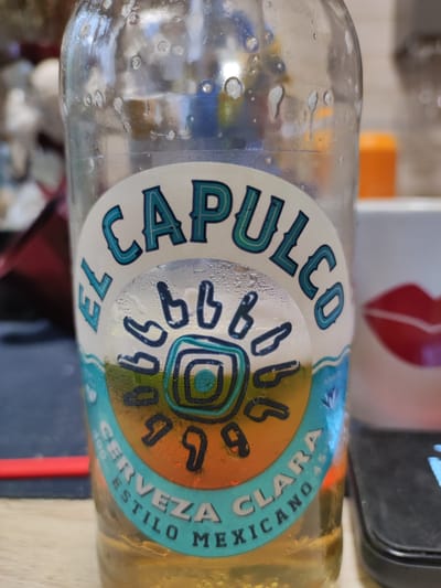 The image shows a close-up of a partially full glass bottle of beer, labeled "EL CAPULCO CERVEZA CLARA ESTILO MEXICANO". The bottle is covered in condensation. Behind the bottle, a white mug with a red lip design is visible. An orange object is out of focus in the background. The scene appears to be indoors. There is no indication of the city of Алабино, Russia.