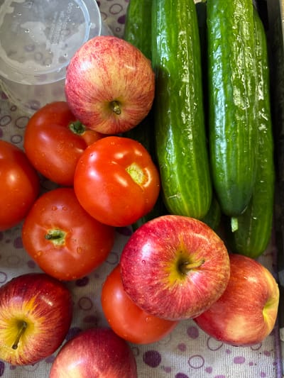 The image depicts a close-up, top-down view of a collection of fresh produce, specifically tomatoes, apples, and cucumbers, arranged on a patterned surface. The produce appears to be recently washed, with visible water droplets on their surfaces.
In the upper left corner, a portion of a clear plastic container is visible, possibly indicating storage or preparation. To its right, an apple with red and yellow streaks is positioned. Below this apple, several bright red tomatoes are clustered. Further to the right, a row of green cucumbers, glistening with water, dominates the upper half of the frame. In the lower half of the image, more apples and tomatoes are arranged, overlapping each other. The patterned surface beneath the produce features circular motifs in shades of purple.
No specific location within Zheleznodorozhnyi, Russia, can be identified from the visual information provided in the image. The scene is forensic in its presentation of objects without any contextual clues to a specific place in that city.
