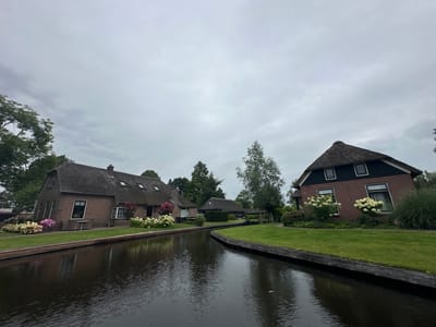 The image shows a picturesque canal in Giethoorn, Netherlands, a village renowned for its waterways and lack of roads. The canal is dark and still, reflecting the overcast sky and the surrounding greenery. On the left side of the canal, a traditional thatched-roof house made of brick is visible. It has several windows and a large thatched roof. In front of the house, a lush green lawn is dotted with blooming hydrangeas in shades of pink and white.
To the right of the canal, another brick house with a thatched roof is prominently featured. This house also has a well-maintained lawn and is adorned with white hydrangeas. Further back, additional houses and trees are visible, contributing to the serene and idyllic atmosphere of the village. The sky is a muted gray, suggesting a cloudy day, but the overall scene is vibrant due to the greenery and flowers. There are no people or boats visible in the image.