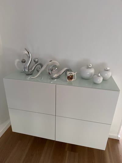This image depicts a modern, white dresser with a glass top, situated in what appears to be a living space in Oberleichtersbach, Germany. The dresser is adorned with several decorative items. On the left side of the dresser top, there are abstract, silver-colored sculptures. In the center, there is a small, white ceramic mug decorated with a picture of a building. To the right, three white, apple-shaped decorative objects with silver stems are arranged. The dresser itself is minimalist, with clean lines and no visible handles, suggesting a contemporary design. The background is a plain white wall. The floor is covered with dark wooden laminate flooring. There are no people visible in the image.