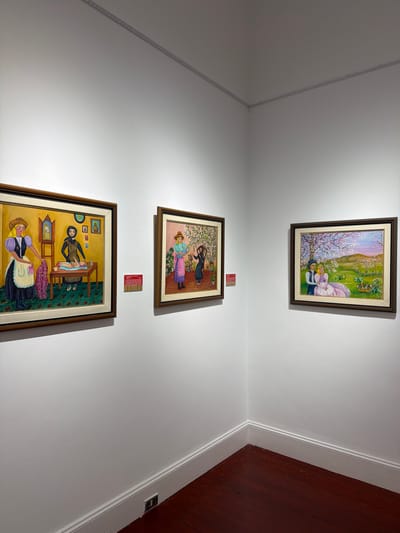 Three framed paintings are displayed on a white wall in a corner of a room with dark red hardwood floors. The painting on the left depicts two women in traditional attire in an indoor setting, with one woman tending to a baby on a table. The center painting shows two women in a garden with patterned wallpaper. The painting on the right portrays a couple sitting in a blooming tree-lined landscape. Red labels are attached to the wall below the left and center paintings. A light fixture runs along the ceiling in the corner.