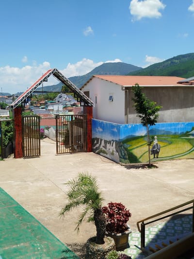 The image is an outdoor scene in the city of K'Rèn, Vietnam, featuring a courtyard area with a prominent entrance gate and a mural on a wall.

In the foreground, a paved courtyard dominates the lower portion of the image. To the left, a green-colored area with red markings suggests a sports court or a similar designated space. A small palm tree is planted in the middle ground to the right, with a reddish-leaved plant in a square pot nearby. A set of stairs with a metal railing is visible in the far right, leading upwards.

The central focus is a large, open gate with a triangular, open-frame roof structure. The gate itself is made of metal bars and is flanked by posts with a wood-grain pattern. Beyond the gate, a street lined with houses and buildings can be seen, indicating a residential area.

To the right of the gate, a long, low wall displays a colorful mural. The mural depicts a rural landscape with rice paddies, a person riding a water buffalo, and buildings in the distance. A small tree is planted in front of the mural, partially obscuring the painting.

In the background, rolling green hills and mountains are visible under a bright blue sky with scattered white clouds. The architecture of the surrounding buildings appears to be of a typical Vietnamese style, with tiled roofs. The overall impression is that of a sunny day in a semi-urban or rural setting. No people are clearly visible within the frame.