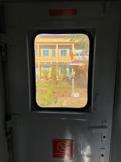 The image depicts the interior of a train carriage, looking out through a window at a building and vegetation. The window is rectangular with rounded corners and framed by dark material. Outside, a yellow building with blue-framed windows is visible. There are trees and bushes in front of the building, and a chain-link fence. The interior of the train carriage is dimly lit and appears to be made of gray metal. A red sign with text in Vietnamese and English, reading "KHÔNG ĐƯỢC ĐỨNG Ở ĐÂY" and "DON'T STAND HERE", is affixed to the door. The location is specified as Châu Ổ, Vietnam.