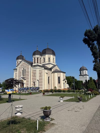 The image shows the exterior of a large, ornate church with multiple domes, a bell tower, and a clear blue sky. The church is painted in a cream color with horizontal reddish-brown stripes. It has several arched windows and crosses atop its domes. In front of the church, there is a paved walkway lined with planters and some greenery. Several benches are visible, as are some framed portraits displayed on a board. A large tree with dark foliage is situated to the left of the church. In the background, to the right of the church, a tall, slender bell tower is visible, also topped with a dome and a cross. The overall environment appears to be well-maintained and serene, suggesting a public space. The location is identified as the city of Mamaivtsi, Ukraine.