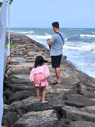 The image depicts an adult male and a child on a rocky embankment adjacent to the ocean in Jembrana, Indonesia. In the foreground on the far left, two vertical strips of white fabric hang. A sloped structure composed of large, irregular dark grey rocks forms the ground, with small patches of green vegetation growing between some stones on the upper left.
A child, viewed from behind, stands barefoot on the rocks in the mid-foreground to the left. She wears a pink long-sleeved cropped top and matching pink shorts. Her long, dark hair is loose and appears to be slightly blown to her right. A dark band is visible on her left wrist.
Behind and to the right of the child, an adult male stands on the rocks, also viewed from behind. He wears a light grey short-sleeved t-shirt, dark shorts, and black sandals. A dark strap, possibly from a bag or camera, crosses his right shoulder, and his hands are positioned as if operating a device in front of him. His dark hair is short and styled.
The turbulent ocean dominates the background and middle ground, with multiple lines of white-capped waves breaking towards the shore. White foam is visible where the waves collide with the rocks. The water exhibits a deep blue-green hue. Above the ocean, a clear, light blue sky extends to the horizon. A small, bright green object rests on a rock in the lower-middle left, near the child's feet.