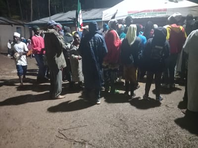 The scene depicts an outdoor nighttime gathering in Butali Chegulo ward, Kenya. Approximately 20-30 individuals, predominantly adults, are standing on a wet, muddy ground surface, casting distinct shadows from an unseen light source. In the mid-ground right, a white canopy tent displays red text identifying "NAMANAMALI COMMUNITY WELFARE CBO" along with a paybill number (400200), account number (58598), and contact information (0727710542). Adjacent to the tent, a flag with vertical green, white, and red stripes is partially visible on a pole. In the left background, simple structures with corrugated iron roofs are present. People are dressed in various casual attire, including jackets, t-shirts, and head coverings such as kofia and headscarves. One individual on the far left holds a small drum-like object. No specific actions beyond standing and mingling are discernible. Dense foliage or trees are visible in the distant background.