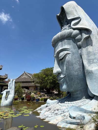 The image depicts a large, weathered, blue-toned statue of a serene figure, likely a deity, situated outdoors in what appears to be a temple complex or garden. The statue is massive, with intricate details on its face, headdress, and clothing. In the foreground, a large, sculpted hand emerges from a body of water filled with lily pads and a single pink lotus flower. The water is dark and reflective. Behind the statue and water, traditional Vietnamese architecture is visible, characterized by dark tiled roofs with decorative elements. Lush green trees and potted plants surround the structures, adding to the tranquil atmosphere. The sky above is a clear, bright blue with a few wispy clouds, indicating a sunny day. The location is identified as Hòa Đa, Vietnam. There are no people visible in the image. The scene is a static, religious or cultural landmark, emphasizing the grandeur of the statue and the surrounding architecture.