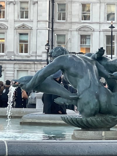 The image depicts a bronze fountain statue in Trafalgar Square, City of Westminster, United Kingdom. The street sign "CHARING CROSS ENT" is visible on a building in the background, confirming the precise location.
In the foreground, the back and left side of a dark bronze statue of a muscular, nude male figure, possibly a Triton or merman, is visible. The figure is holding a dolphin in its extended left arm, from which a steady stream of water erupts vertically into the lower basin of the fountain. The statue's surface shows evidence of water runoff, consistent with its function. The statue is positioned within a multi-tiered stone fountain structure, with light-blue water visible in the basin, reflecting ambient light.
In the mid-ground and background, several indistinct individuals are present, appearing as pedestrians or visitors to the square. One person to the left wears dark clothing and possibly a backpack. Another person, positioned behind the fountain, wears a dark beanie and dark clothing, with a dark beard. The individuals are largely static or subtly moving, with no discernible specific actions or direct interactions captured.
The background consists of light-colored, multi-story stone buildings featuring numerous rectangular windows with white frames, indicative of classical urban architecture. A black lamppost with an ornate top is visible between the statue and the buildings. The lighting suggests daytime or early evening conditions.