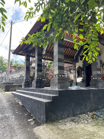 The image presents an upward perspective of a Balinese Hindu temple or shrine in Pejukutan, Indonesia. The structure features a traditional design with dark gray, vertically striped pillars supporting a wooden roof. Ornate carvings decorate the upper sections. A lush green tree branch with leaves is visible on the left, partially obscuring the view. The background showcases the surrounding temple complex with similar architectural elements and glimpses of palm trees and other vegetation. A concrete staircase leads up to the structure. The ground shows signs of wear and debris. An electrical pole with wires is visible against a backdrop of a cloudy sky.