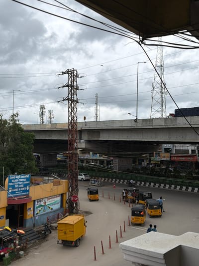 The image captures a cloudy day in Shamshabad, India, likely near a major intersection. In the foreground, a yellow auto-rickshaw truck is parked. To its left, a motorcycle is parked near the entrance of a yellow building labeled "SULABH COMPLEX PUBLIC TOILET BATH & URINALS PAY & USE LADIES & GENTS". A mural is painted on the side of this building. Further into the scene, an elevated highway crosses overhead, supported by concrete pillars. Several tall communication towers are visible on either side of the highway. On the road below the highway, a line of yellow auto-rickshaws and a few cars are present. Two men are interacting near one of the auto-rickshaws. The sky is overcast with heavy clouds, and electrical wires crisscross the upper portion of the frame.