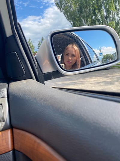 This is a medium-shot, close-up photograph taken from inside a car, looking at the side-view mirror. The mirror reflects a young woman with blonde hair, looking directly at the camera with a neutral expression. She is inside the car.

The background of the mirror reflection shows a street scene with a pedestrian crossing sign, trees, buildings, and a bright blue sky with white clouds, indicating it is daytime and fair weather. In the mirror, the woman's face is in focus, with her head tilted slightly. A smartphone is visible in her hand, also reflected in the mirror.

The foreground of the image, outside the mirror, is dominated by the interior of the car. The door panel is visible, with a speaker grille, and a section of faux wood trim. The car's door frame and a part of the window are also visible. The overall lighting suggests it is a sunny day. The location is identified as Chulki-Sokolovo, Russia. No visible text is present in the image other than on the pedestrian crossing sign in the reflection.