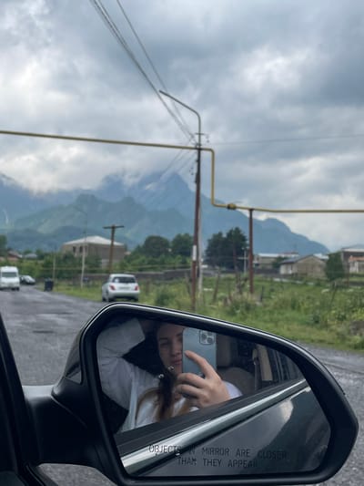 A reflection in a car's side mirror shows a young woman taking a selfie with a smartphone. She is wearing a white shirt and has her left hand near her head. The mirror also reflects the surrounding environment, which appears to be a rural road in Garbani, Georgia. The road is wet, suggesting recent rain. In the background, there are mountains under a cloudy sky, with scattered houses and trees visible. A white van and a silver car are on the road. Power lines and poles are also present in the scene. The text "OBJECTS IN MIRROR ARE CLOSER THAN THEY APPEAR" is visible at the bottom of the mirror.