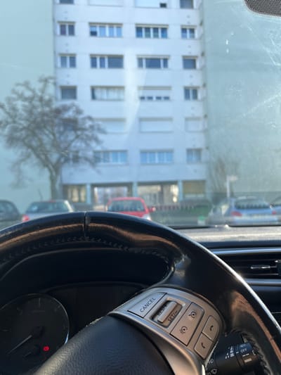 The image depicts a view from the interior of a vehicle, looking forward through a smudged windshield. In the immediate foreground, a black, leather-wrapped steering wheel with visible stitching occupies the lower portion of the frame. The right spoke of the steering wheel features a silver control panel with buttons labeled "CANCEL," "RES/+", and "SET/-," along with additional symbols for cruise control, audio, and phone functions. Part of the vehicle's instrument cluster, showing a speedometer or tachometer gauge with a lit red circular warning indicator, is visible to the lower left. A segment of the dashboard and a wiper stalk are also discernible.
Beyond the vehicle's interior, the scene unfolds into an urban residential environment under bright daylight, with sunlight creating reflections and streaks on the windshield. Several vehicles are parked on a paved surface, likely a street or parking lot. A red hatchback is prominently positioned directly ahead, partially obscured by the steering wheel. To its left, a darker-colored car is visible, and to its right, a light blue or silver hatchback is parked.
In the background, a large, multi-story residential apartment building dominates the view. The building is light-colored, likely white or light grey, and features numerous rectangular windows arranged symmetrically across its facade, some equipped with external white roller shutters. The ground floor exhibits a different architectural treatment, with larger window openings, possibly indicating common areas or commercial spaces. To the left of the building, a bare deciduous tree with a visible trunk and branching structure stands without foliage.
The location is identified as the **Val Saint-Clair district, specifically near Boulevard des Belles Portes, in Hérouville-Saint-Clair, France.**