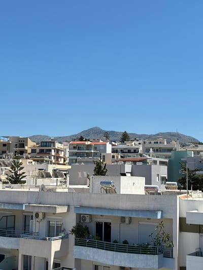 The image shows a view of buildings with mountains in the background under a clear blue sky. The buildings are a mix of white and pastel colors, with balconies, windows, and air conditioning units visible. Several solar water heaters are mounted on the rooftops. The style of the architecture suggests a Mediterranean location. The presence of solar water heaters is common in Greece, and the architectural style is consistent with that found in cities like Rethymno, Greece.