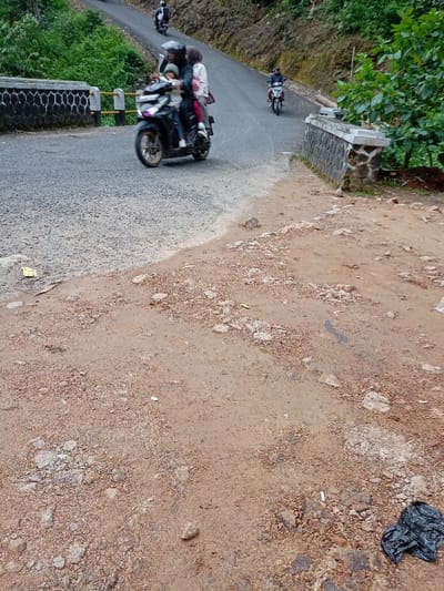 The image shows a dirt road with a motorcycle and a couple of people on it. The road appears to be in poor condition, with loose gravel and dirt. In the background, there is a stone wall, lush foliage, and a paved road. The motorcycle is a black scooter with a man and two women on it. They are all wearing helmets. The man is driving the motorcycle. The two women are sitting behind him. One of the women is holding a child. There is a second person on a motorcycle in the background. The motorcycle is being driven by a man in a black jacket and black helmet. The image appears to have been taken in Cisewu, Indonesia, at the coordinates -7.41, 107.541.