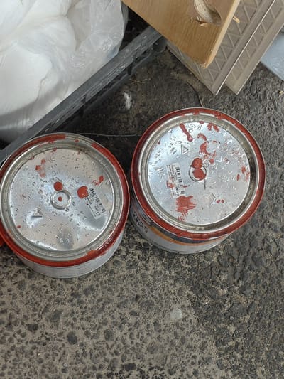 A high-angle shot captures two cylindrical metal cans, heavily stained with red paint or primer, resting on a dark, coarse asphalt or concrete surface. The cans are viewed from their tops, revealing silver lids splattered with red residue and covered in condensation or water droplets.
Both cans feature barcodes and partially legible text in Cyrillic. The top can's lid displays "ГРУНТОВКА" (Primer) and an EAN code "4 699566 979832". The bottom can's lid shows an EAN code "4 690566 979910", along with "FORMULA OS" and "партия: 5240" (batch: 5240). The red stains on the lids and rims indicate the cans have been used or have leaked.
The immediate surroundings suggest a construction or renovation environment. To the lower left, a large, translucent white plastic bag, likely containing powdered building material, is visible. Adjacent to this, pieces of light-colored wooden debris and fragments of what appear to be concrete tiles or textured building panels are present. A dark, possibly metal, frame or grate also runs along the left side.
The setting is an outdoor or semi-outdoor location, likely a worksite or storage area in Nadym, Russia. The soft, diffused lighting and the presence of water droplets on the cans suggest damp or overcast weather conditions. No people or active events are depicted; the scene shows discarded or temporarily stored materials.