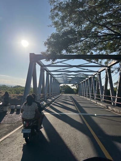 In Kubu, Indonesia, two individuals are riding a scooter across a metal truss bridge on a sunny day. The sun is bright, casting long shadows from the bridge's structure onto the asphalt road. The scooter has a license plate that reads "DK 3171 ADO". Trees line the sides of the road leading up to and over the bridge, with lush green foliage visible in the background. The bridge itself is a prominent feature, with its metallic beams creating a geometric pattern against the sky. The sky is clear and blue, with a few wispy clouds in the distance. The overall scene depicts a serene journey across a bridge in a tropical environment.