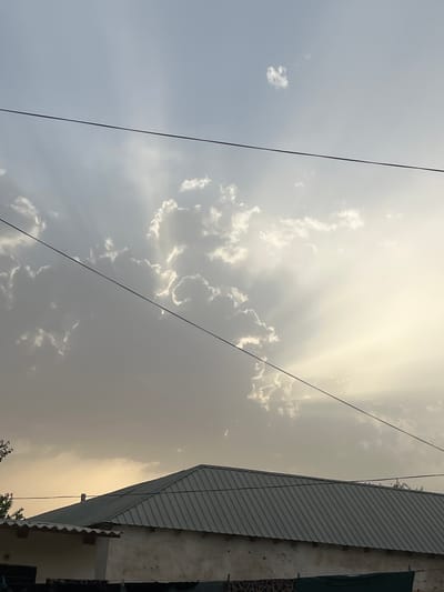 The image depicts an outdoor scene with a focus on the sky and a building. The sky is filled with clouds, some of which are illuminated by crepuscular rays, suggesting it is either sunrise or sunset. The light is warm and hazy, indicating a specific time of day and possibly a weather condition like dust or humidity. In the foreground, two dark, thin wires run diagonally across the frame, obscuring parts of the sky.
On the right side of the image, a portion of a building is visible. It has a sloped, corrugated metal roof and a rough, textured wall. The building appears to be in a somewhat weathered state. Some foliage is also visible at the bottom of the frame, hinting at an outdoor environment.
The overall impression is a somewhat dramatic sky with the sun's rays breaking through the clouds, juxtaposed with the mundane elements of utility wires and a simple building. There is no visible text. The scene evokes a sense of calm or perhaps a transient moment of atmospheric beauty. The provided location context of Balkh, Tajikistan, suggests a setting in a region that experiences distinct weather patterns and has a particular architectural style.