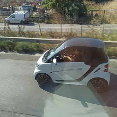 A white smart car is driving on a road in Municipal Unit of Echedoros, Greece. The car has a black roof and a person wearing glasses is driving it. The car's windows are tinted. In the background, there is a white van, a fence, and trees. The road appears to be a highway or main road, with a concrete barrier on the side. The image is taken from another moving vehicle, as evidenced by the motion blur.