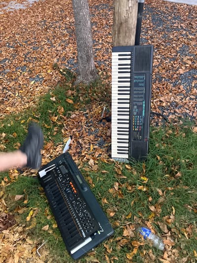 The image presents a still life scene, possibly outdoors, in Saladillo, Argentina. Two musical keyboards are placed on a ground covered partially with gravel and dead leaves, and partially with green grass and dead leaves. The top keyboard, a Kawai FS650, lies partially on the gravel and leaves, and partially on the grass and leaves. A log is also located between the gravel/leaves, and the grass/leaves. An empty plastic water bottle is visible on the grass and leaves to the upper right of the frame. Below the Kawai FS650, an Arturia MiniFreak keyboard rests on a box on the grass and leaves. A foot and a part of leg are captured at the bottom of the frame.