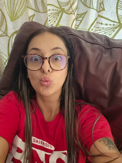 A close-up selfie shows a person lying down, with their head turned towards the camera. They are wearing a red t-shirt with a white logo that reads "SMIRNOFF." The person has long, dark hair and is wearing glasses with a tortoiseshell pattern. Their lips are pursed in a kissing gesture, and they have a small hoop nose ring. The background consists of a brown textured surface, likely a couch, and a curtain with a green leaf pattern. The lighting appears to be natural, suggesting it might be daytime. The overall mood is casual and playful. There is no specific indication of time of day, weather, or a precise location within Bridlington, United Kingdom, beyond what can be inferred from the domestic setting.
