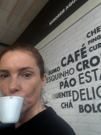 An indoor, eye-level shot captures a person, likely female, drinking from a white cup, possibly Styrofoam, with a beverage inside. She has brown hair pulled back, and is wearing a black long-sleeved top. Her visible accessories include small hoop earrings and a silver cross pendant necklace.
The background is a white wall with a textured, brick-like pattern, adorned with large black Portuguese text, suggesting a menu or slogan. Above this, a black horizontal strip also displays white text. The image is oriented diagonally, with the person's head tilted to the right.
Visible text on the black strip reads "SABOREIE AQU" (likely "SABOREIE AQUI" - "Enjoy here"). On the white wall, fragmented words are discernible, indicative of a cafe or snack bar menu: "CAFÉ" (coffee), "SUMO" (juice), "PÃO" (bread), "QUENTE" (hot), "CHÁ" (tea), and "BOLO" (cake). Other fragments include "CHE", "PING", "CUR", "ESQUINHO", "CRO" (likely part of "Croissant"), "EST", and "DELIC" (likely part of "delicioso" - delicious).
The setting appears to be a casual eatery, possibly in Cacém e São Marcos, Portugal, given the language and context. The person is engaged in the simple activity of drinking, with a neutral expression. No specific time of day or weather is indicated.