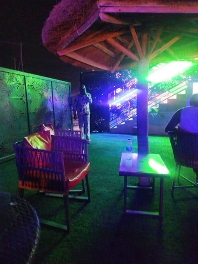 A vibrant, dimly lit outdoor space features green artificial turf. A thatched-roof structure casts shadows over seating. A man in a patterned shirt stands near a green decorative partition and a brightly lit screen displaying geometric designs. Tables hold a water bottle. One chair is draped with a bright yellow cloth, adding a pop of color to the scene.