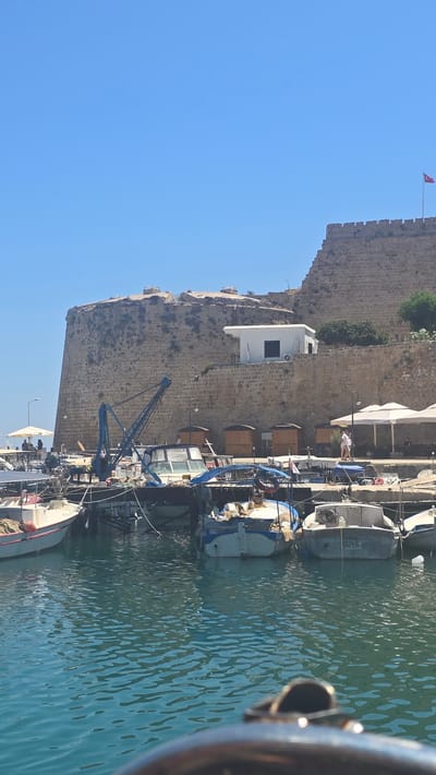 In Kyrenia, Northern Cyprus, a picturesque harbor scene unfolds under a clear blue sky. A stout, ancient stone castle, likely Kyrenia Castle, dominates the upper portion of the frame, its weathered ramparts and battlements a testament to centuries of history. A Turkish flag flutters from a pole atop the castle walls. Below the castle, a bustling harbor is filled with numerous boats of varying sizes, bobbing gently on the turquoise water. Some are small fishing vessels, others appear to be pleasure crafts. A blue crane stands prominently among the boats, suggesting ongoing activity or maintenance. Along the waterfront, several small, wooden kiosks are visible, and white umbrellas shade outdoor seating areas, hinting at a vibrant waterfront promenade with cafes or restaurants. A few people are casually strolling or sitting, enjoying the sunny atmosphere. The water reflects the blue of the sky and the textures of the surrounding structures.