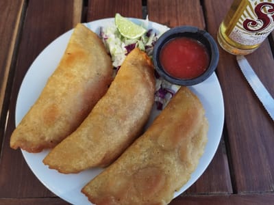 The image shows three golden brown, deep-fried, crescent-shaped pastries on a white plate. The pastries are arranged in a semicircle with one pastry on the left, two pastries on the right, and some lettuce and a red sauce dipping bowl in the center. The pastries are partially covered with lettuce and a red sauce dipping bowl, which contains a red, presumably tomato-based sauce. To the right of the plate, there is a bottle of beer, which has the word "Servizio" in the lower right corner, and a butter knife. The pastries are located in the city of Unidad de Riego, Mexico. The scene is set on a wooden surface.