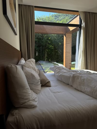 A bedroom scene at sunrise in Bakovići, Montenegro. The room features a large bed with white linens and pillows, facing a large window overlooking a forest and mountains. Natural light streams into the room, illuminating the bed and the wooden architectural elements of the window frame. The curtains are partially drawn, revealing the scenic view outside.
