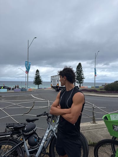 The image depicts a male subject standing alongside a coastal road at Bondi Beach, Australia, under an overcast sky. The man, positioned on the right, faces left in profile, his arms crossed over his chest. He wears a black sleeveless shirt and black shorts, with white wired earphones visible around his neck. He stands next to a grey "PEDAL" branded bicycle; a black helmet with a white checkered pattern and another dark helmet are secured to its rear rack. Part of a green bicycle rental basket is visible on the far right.

In the midground, a paved road stretches horizontally, separated from the bicycles by a wavy metal barrier. Beyond the road, the scene includes the ocean with visible waves, and several indistinct figures near the water. A white truck with a container is driving away from the viewer on the road, with other vehicles partially visible. Two tall streetlights, each featuring a blue banner, punctuate the landscape, alongside two prominent conifer trees and areas of green landscaping. Colorful concrete barriers are arranged along the road's edge. The background is dominated by the grey, cloudy sky meeting the ocean horizon.