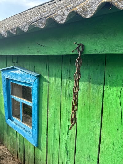 The image shows the exterior of a weathered, green wooden structure, likely a shed or outbuilding, located in the village of Chuy-Atasevo, Russia. A small, bright blue window with two panes is visible on the left side of the structure. Above the window, a rusty chain hangs from two nails driven into the wooden wall. The roof of the structure is covered with corrugated asbestos-cement sheets, appearing aged and possibly covered in lichen. The wood of the walls is vertically oriented and shows signs of wear and cracking. The ground in front of the structure appears to be dirt and dry vegetation. The lighting suggests it is daytime, with shadows cast on the green wall.