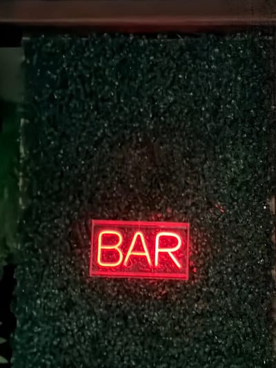 A red neon sign that spells "BAR" is mounted on a dark green textured wall. The sign is centered horizontally and located in the lower third of the image. The wall appears to be made of artificial greenery. Above the wall, a darker, possibly wooden, surface is visible. The lighting is dim, emphasizing the glow of the neon sign. The image is a close-up shot, focusing on the sign and the wall behind it. The location of this scene is identified as Orhei, Moldova.