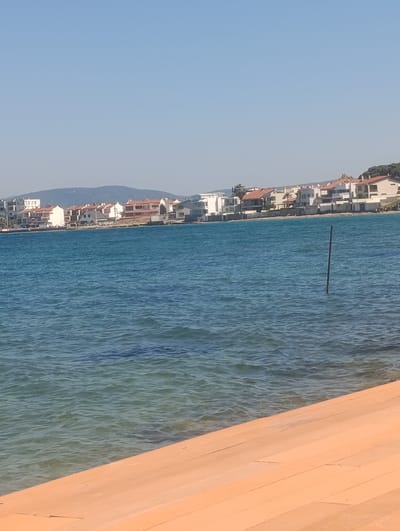 The image depicts a serene waterfront scene in Urla, Turkey, under a clear blue sky, suggesting a sunny day. In the foreground, an orange wooden deck or boardwalk occupies the lower right portion of the frame, providing a warm, inviting texture. Beyond the deck, the calm, turquoise water of the sea stretches towards a distant shoreline.

The middle ground features a line of houses with terracotta roofs and white facades, characteristic of Mediterranean architecture. These buildings are nestled along the coast, with some showing signs of vegetation, including palm trees. A thin, dark pole stands vertically in the water, emerging from the gentle ripples. In the background, a hazy mountain range forms a soft silhouette against the horizon.

The overall impression is one of tranquility and peacefulness, with the bright sunlight and clear water evoking a sense of a pleasant, warm day. There are no people or animals visible, emphasizing the quiet solitude of the location. The scene appears to be captured during daylight hours, likely late morning or early afternoon, given the brightness and lack of shadows. No visible text is present in the image.