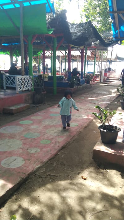 A young child, wearing a light blue hooded jacket and grey pants, walks on a patterned pink and circular paved walkway in Reudeuep, Indonesia. The walkway is bordered by dirt and a raised concrete curb with potted plants. Behind the child are colorful open-air structures with thatched roofs, supported by brightly painted poles. Several people are visible in the background, some seated under the structures. The environment appears to be an outdoor seating area, possibly near a beach or park, with sunlight filtering through trees, casting shadows on the ground.