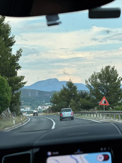 This is a vertical, eye-level shot of a scenic drive along a winding road, likely in Seget Donji, Croatia, based on the provided location context. The perspective is from inside a vehicle, looking out through the windshield and side window, suggesting a road trip or journey.
The main subjects are the road itself, which curves and slopes downwards to the right, bordered by a guardrail. Trees and lush green foliage line the roadside, with a distant view of mountains and a village nestled on the hillside. A silver hatchback is visible on the road ahead, and another dark vehicle is further down.
A prominent triangular road sign with a red border and a black symbol indicating falling rocks or debris is positioned on the left side of the road, warning drivers of potential hazards. A smartphone displaying a navigation app is visible on the right side of the frame, indicating the use of GPS for guidance. The sky is partly cloudy, with streaks of white clouds against a pale blue backdrop. The lighting suggests it's daytime, possibly late afternoon or early morning, with soft light illuminating the landscape. There are some visible specks on the lens or glass, which appear as small white dots throughout the image.