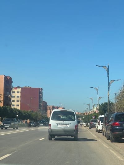 The image captures an urban street scene in Beni Mellal, Morocco, on a clear, bright day. The perspective is from inside a vehicle, looking forward along a multi-lane asphalt road. Directly in the foreground, centered in the frame, is the rear of a light-colored Peugeot Partner van with Moroccan license plate "154817 | 61".
To the left of the Peugeot van, in an adjacent lane, a black sedan is visible, followed by other vehicles further down the road. To the right, a paved shoulder or parking area contains several parked vehicles, including a dark-colored SUV (possibly a Porsche Cayenne), a white van, and another dark SUV, all facing away from the camera.
Along the right side of the road, a series of modern streetlights with distinctive ornate, leaf-shaped fixtures are mounted on poles. Beyond these lights, multi-story buildings with reddish-brown and light-colored facades line the street. Similar residential or commercial buildings, predominantly terracotta in hue, are visible along the left side of the street, beyond a strip of trees and shrubs.
The sky above is a uniform, clear blue, indicating sunny weather. Further down the road, a motorcycle is visible in the distance, navigating traffic. The scene depicts typical daytime urban traffic flow and infrastructure in a Moroccan city.