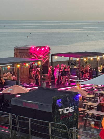 This image captures an outdoor event with a tropical theme, likely in Brighton, United Kingdom, given the context clues of the "TIDE" signage and the beach setting. The scene is set at dusk, with the sky displaying a soft gradient of grey and a hint of pale blue. The calm sea stretches to the horizon, with a few small buoys visible in the water.
In the foreground, a dark-colored structure with "SHISHA" illuminated in pink neon lettering runs along its top edge. Adjacent to this is a section of artificial greenery with "TIDE" prominently displayed in white block letters, accompanied by smaller text below it. This area is lit by blue neon lights. Beyond this, a series of wooden picnic tables and benches are arranged on a wet, reflective surface, suggesting recent rain or a cleaning.
The central focus of the image is a raised, open-air stage area, adorned with vibrant pink neon lights. A large, tiki-style structure with a carved face and a roof featuring the same pink neon lighting dominates the backdrop of this stage. Several people, mostly women dressed in evening wear, are gathered on and around the stage, some appearing to be dancing or socializing. One individual in a white hat and a light-colored dress stands out. String lights with colorful triangular flags are strung across a covered seating area to the left of the stage, where other individuals are seated and interacting. An orange umbrella provides shade over a section of this seating area. The overall atmosphere suggests a lively event or party taking place at a beachside venue.
