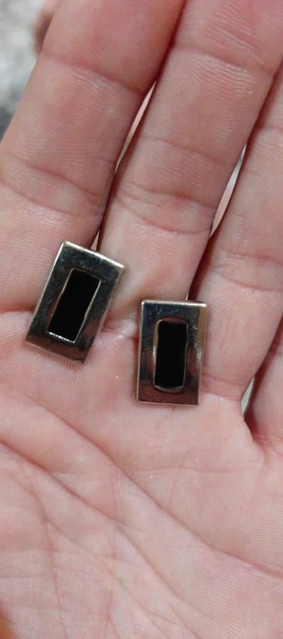 A close-up, overhead shot shows two silver-colored rectangular cufflinks with black inserts lying on a person's hand. The cufflinks are positioned with their longest sides horizontal. The background of the image is blurred and appears to be a light-colored, textured surface. The location of this image is not specified within the image itself, and it is not possible to determine the city of Kalininskaya, Russia from the visual information provided.