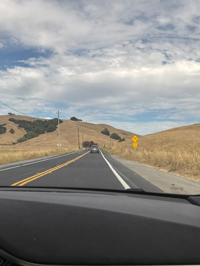 The image depicts a view from the interior of a vehicle traveling on a rural road, likely in or near Petaluma, California, United States, given the characteristic golden hills and cloudy sky. The road has a black asphalt surface with double yellow lines in the center and a white line on the right edge. In the foreground, the dark dashboard of the vehicle is visible. Further down the road, several cars are traveling in the same direction, with the closest one appearing to be a silver SUV. On the right side of the road, a yellow diamond-shaped sign with a bicycle symbol and the text "SHARE THE ROAD" is mounted on a pole. The landscape flanking the road is characterized by dry, golden-brown hills covered in grass and scattered clusters of green trees. The sky above is a mix of blue patches and white, fluffy clouds. Power lines are visible stretching across the sky.