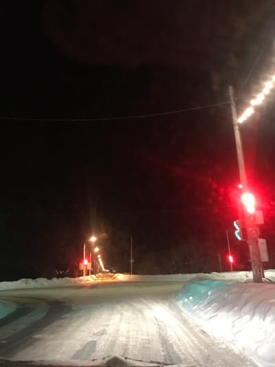 The image displays a nighttime winter street scene in the city of Kremyonki, Russia, viewed from a low perspective, likely from inside a vehicle. The foreground reveals a snow-covered asphalt road with distinct tire tracks, flanked by elevated snowbanks. To the right, a utility pole stands prominently, supporting a traffic light emitting a bright red signal. A fainter green signal is visible beneath the red light. A string of luminous white lights is wrapped around the upper section of this utility pole, extending vertically. Another red traffic light is observable further down the road on the left. The road continues into the distance, lined by a series of streetlights on utility poles that cast an amber glow on the snowy environment. The background is dominated by a dark, featureless night sky, with subtle, distant outlines of terrain or structures visible on the horizon. A thin wire traverses horizontally across the upper-mid portion of the frame. No people or active interactions are depicted.