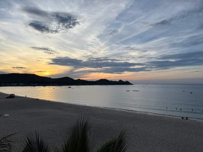 This is a landscape photograph of a beach in Song Cau, Vietnam, taken at sunset or sunrise. The sky is filled with dramatic clouds, streaked with hues of orange, yellow, and blue, with darker gray clouds scattered throughout. The sun is low on the horizon, casting a warm glow across the water.
The foreground consists of a wide expanse of sandy beach. In the mid-ground, the calm ocean meets the shore, with gentle waves lapping at the sand. There are several small boats scattered across the water, some closer to shore and others further out. A red truck is parked on the left side of the beach, near the water's edge. A single person is walking along the shoreline near the truck. In the distance, a line of dark, hilly islands forms the horizon.
In the far right mid-ground, several individuals are visible in the water, appearing to be swimming or wading. Closer to the shore on the right side of the image, two people are sitting on the sand. Palm fronds are visible in the bottom corners of the frame, framing the scene. The overall atmosphere is peaceful and serene, capturing the beauty of the coastal landscape at dusk or dawn.