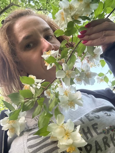 This is a portrait-style, low-angle shot of a person holding jasmine flowers. The person's face is the primary subject, with their eyes wide open and looking directly at the camera. Their blonde hair is visible, framing their face. Their left hand, with bright red nail polish, is reaching into the frame from the top left, gently holding a branch of jasmine flowers. The flowers are white with yellow centers and are in full bloom, with green leaves surrounding them. The background is a mix of the sky and a light gray hoodie worn by the person, which has some text and sequin embellishments. The text on the hoodie is partially visible and appears to be dark gray. The overall impression is one of natural beauty and a candid moment. The image appears to be taken outdoors, given the presence of flowers and the bright, natural lighting, suggesting it might be daytime. The location context of "Дзержинец-83 Снт, Russia" indicates a dacha or garden setting, common in Russia. The composition and the subject's gaze create an intimate and engaging portrait.